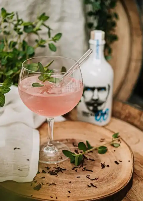 "Glas mit rosafarbenem Gin und frischen Blättern auf einem Holzbrett, im Hintergrund eine Flasche KNUT HANSEN Dry Gin"