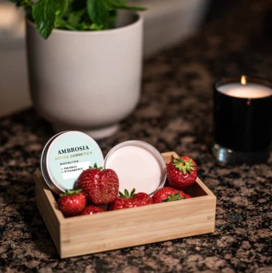 Bodybutter in Dose in einer Holzkiste mit Erdbeeren, daneben Kerze in dunkler, häuslicher Umgebung