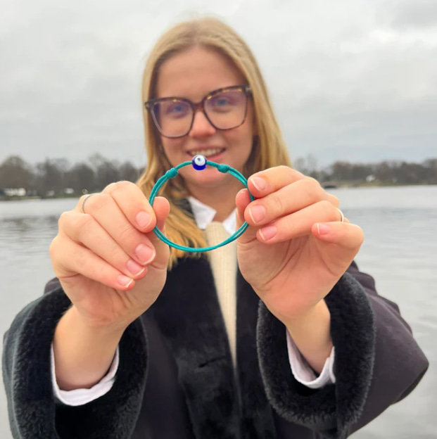 Blonde junge Frau hält ein türkisfarbenes Upcycling-Armband in beiden Händen, See im Hintergrund